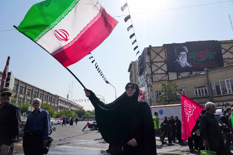 Image: A woman waves an Iranian flag. A banner showing a portrait of the late Supreme Leader Ayatollah Ali Khamenei is seen hanging on a building wall in the background.