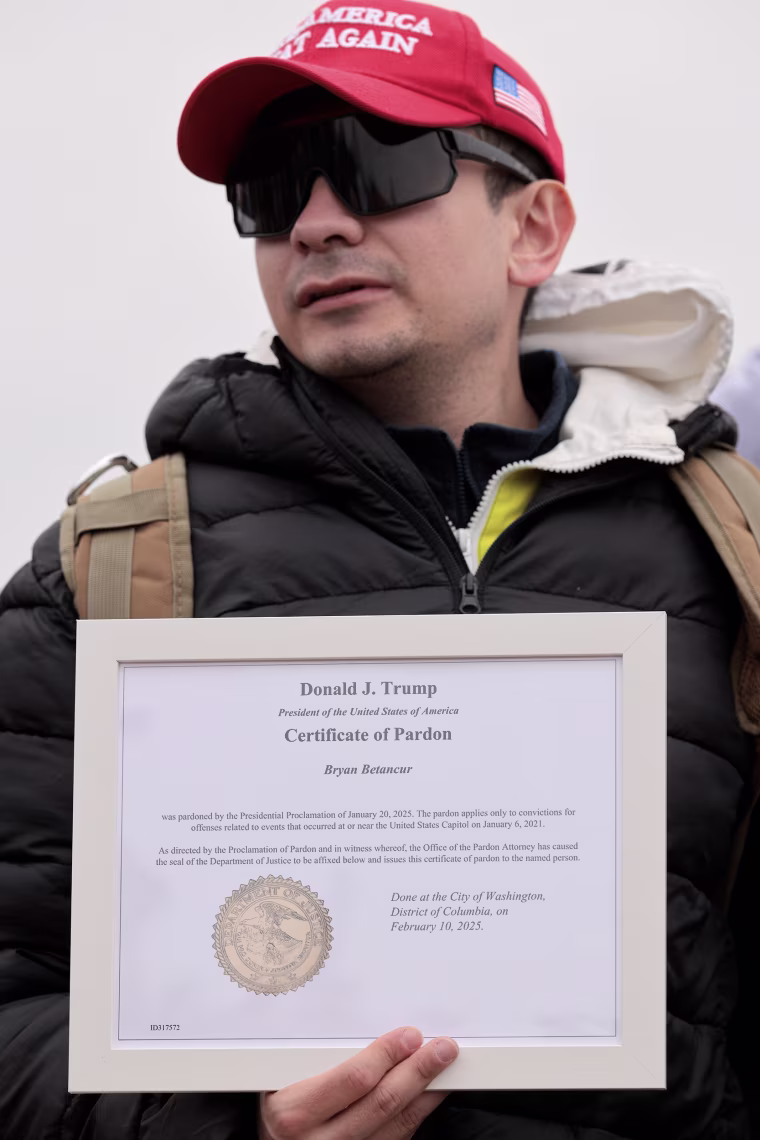 A man wearing a Make America Great Again hat and dark sunglasses holds a framed certificate of pardon from President Trump.