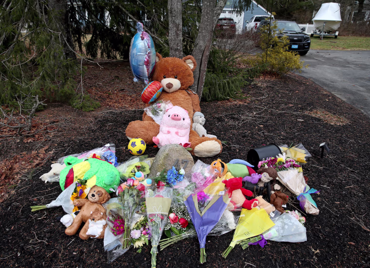 A makeshift memorial of stuffed animals and bouquets lying on the ground next to a residential driveway.