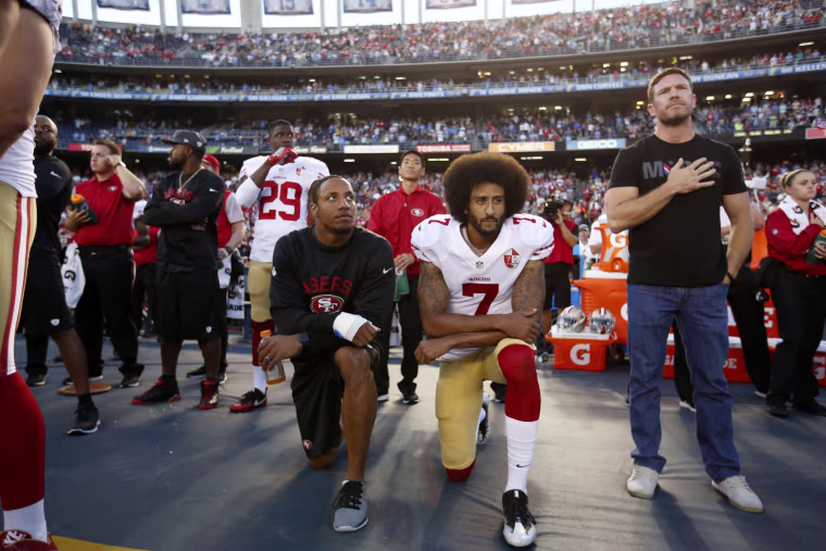 Reid and Kaepernick take a knee on the sidelines of a football stadium while another player holds his hand over his heart.