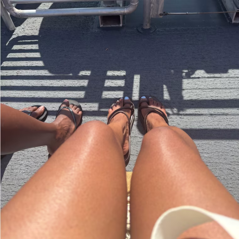 Two feet wearing the Crocs Miami sandals in brown beside two feet wearing plain black flip flops with a grey background.