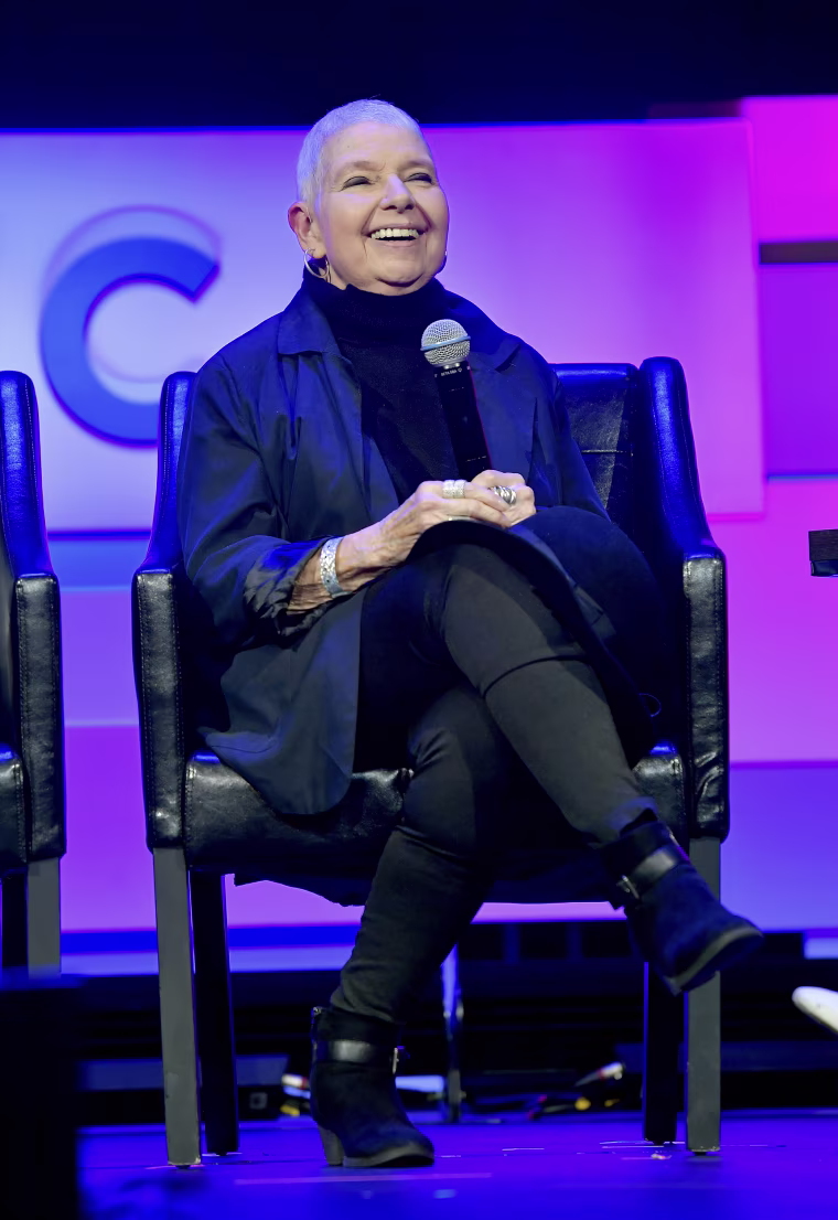 Robin Morgan laughs while seated on stage under blue and magenta lighting.