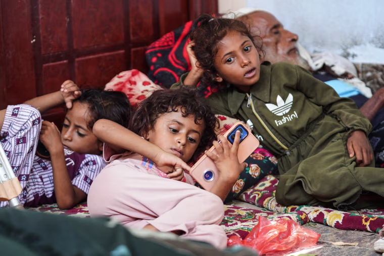 Children lay on the floor on top of blankets, one using a smartphone