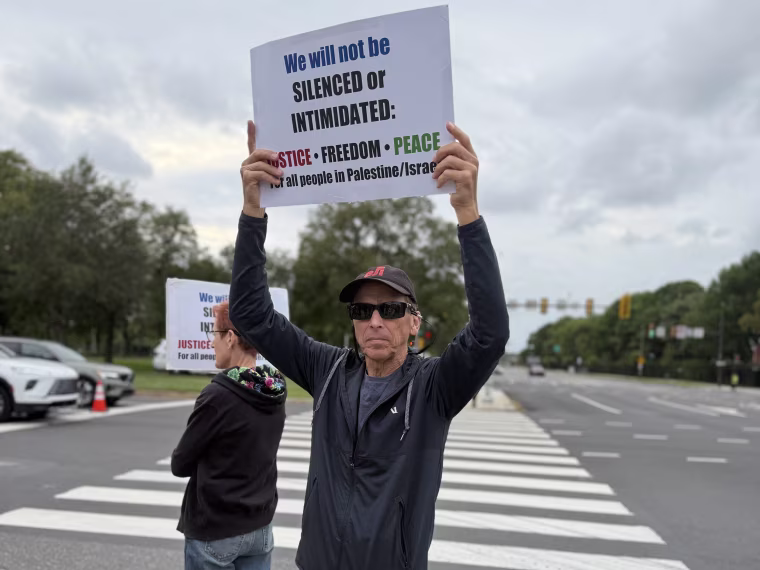 A man in sunglasses holds a sign reading "we will not be silenced or intimidated".