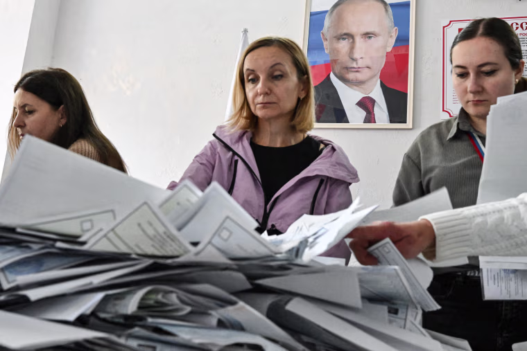 Three people count paper ballots spread atop a table, a framed photo of Vladimir Putin is on the wall behind them