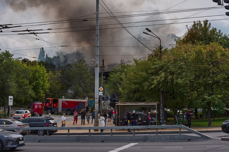 People wait at a public transport stop as a city market burns.