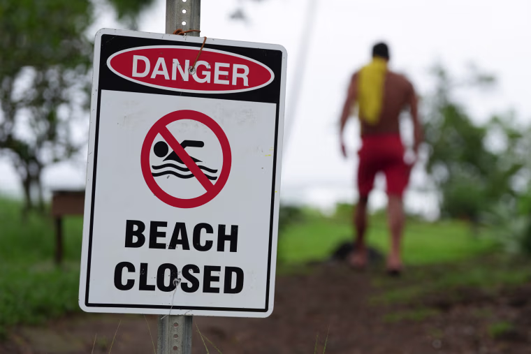 A lifeguard walks back to his tower in the distance, in the foreground in focus is a sign that reads "Danger Beach Closed"