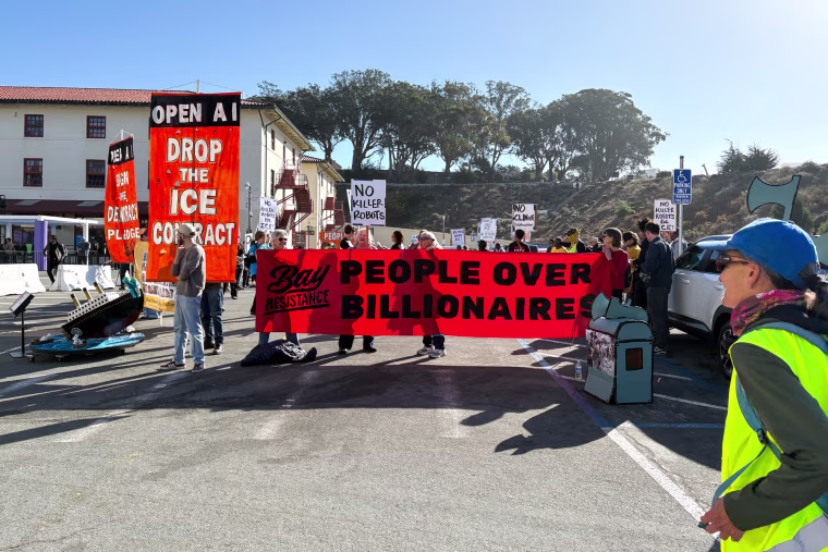 Protestors stand outside of a building holding Anti-Ai posters, a huge banner in the front reads "people over billionaires."