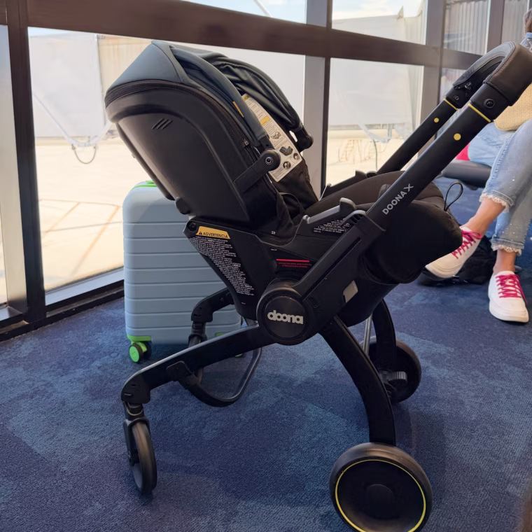 A black Doona X infant car seat in stroller mode, wheels extended, parked on blue carpet by the windows of an airport gate beside a light blue carry-on suitcase.