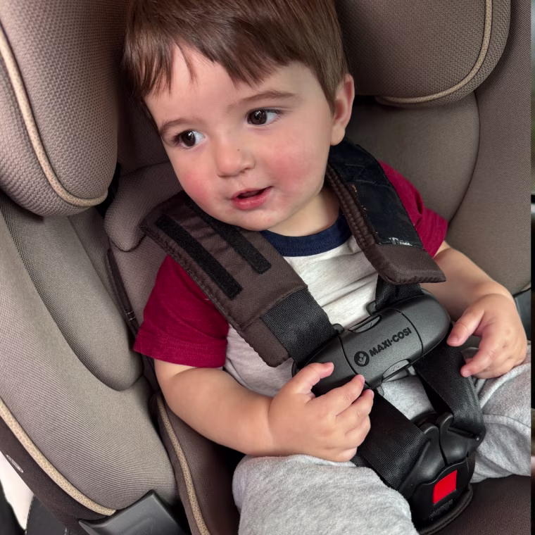 A smiling toddler with brown hair sits in a taupe Maxi-Cosi convertible car seat, buckled into a five-point harness with padded shoulder straps and a Maxi-Cosi-branded chest clip.
