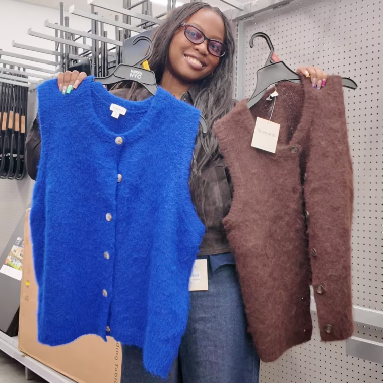 Photo of a woman holding a brown sweater vest and a blue sweater vest on hangers in a store.