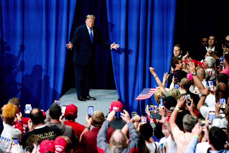 Donald Trump stands in front of an opening through blue curtains on a stage in front of a crowd of supporters
