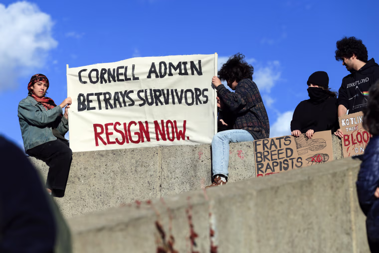 Protestors sitting on a stone staircase hold a sign that reads "Cornell Admin Betrays Survivors, Resign Now"