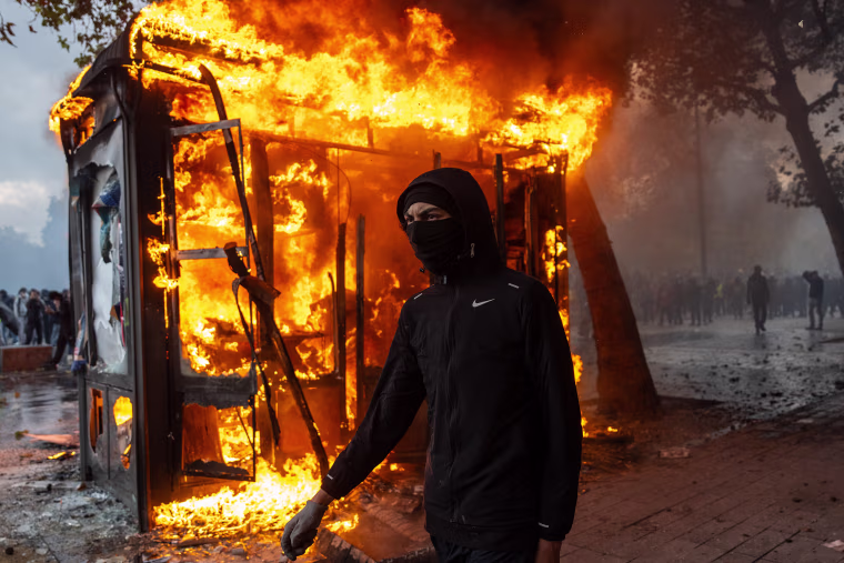 Image: A masked protester walks past a burning news-stand.