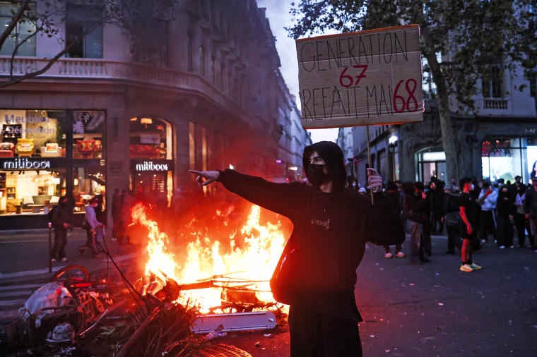Image: A masked protestor holds a cardboard sign reading "Generations 6-7 redoes May 68" in front of a fire at the Boulevard des Capucines.
