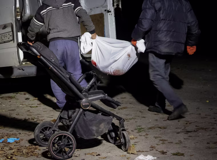 Two men carry the body of a child in a white sheet, with an empty stroller in the foreground of the photo.