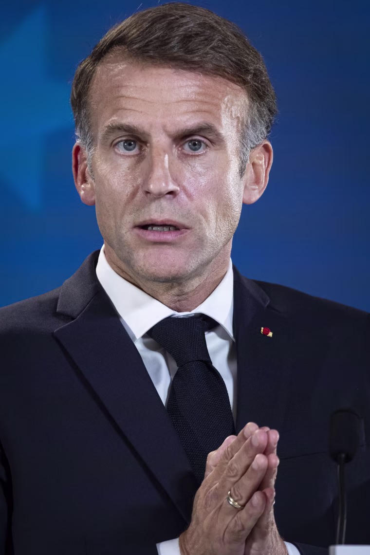 Image: Emmanuel Macron looks on, in front of a blue backdrop.