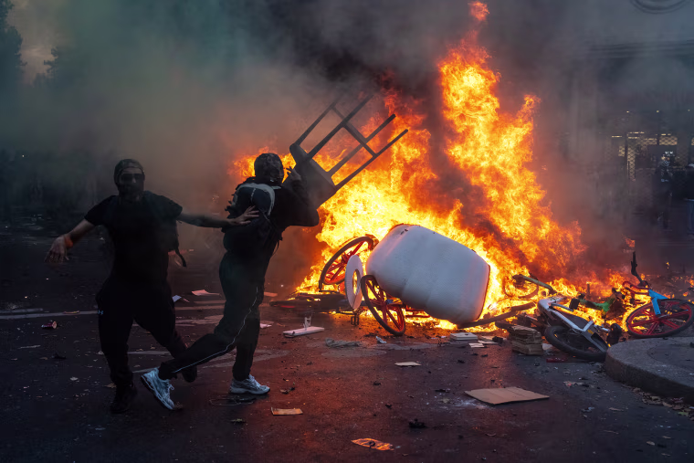 Image: A masked protester throws a chair into a fire in the street.
