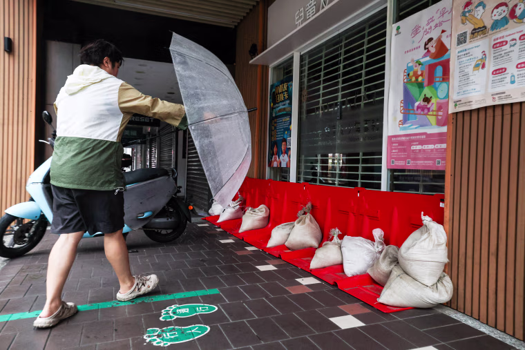 Image: A pedestrian holding an umbrella walks past sandbags and flood barriers placed outside a building.