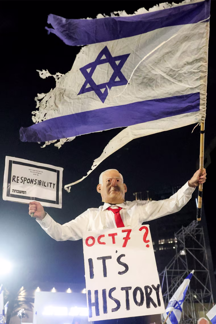 Image: A protester wearing a mask depicting Israeli Prime Minister Benjamin Netanyahu with an elongated nose, evoking the literary character Pinocchio, poses with a placard during an anti-government demonstration.