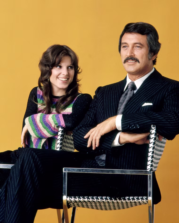 Susan Saint James sitting beside Rock Hudson in front of an orange backdrop