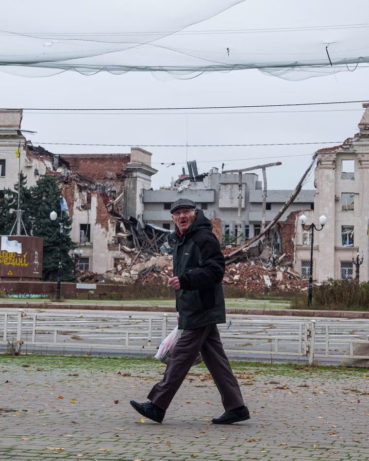 Image: A man walks beneath anti-drone nets stretched over a street in front of a destroyed regional administration building
