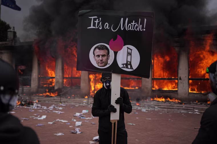 A protester holds a sign with a photograph of French President Emmanuel Macron in front of a burning office, October 17, 2026. 
