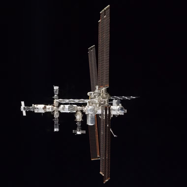 The International Space Station's solar panels are nearly edge-on in this view. Two Russian Soyuz lifeboats and two Progress cargo ships are docked on the left side.