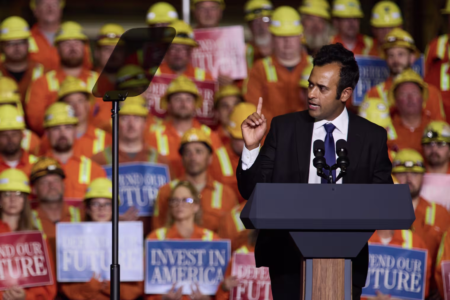 Vivek Ramaswamy speaking at a lectern in front of a riser full of people in orange vests and helmets.