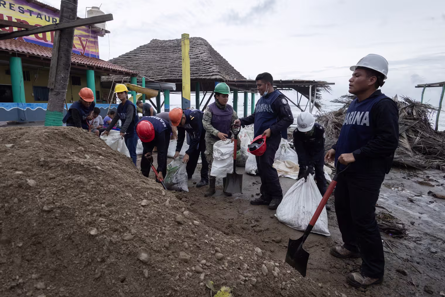 Several people wearing hard hats fill sandbags near a shoreline.