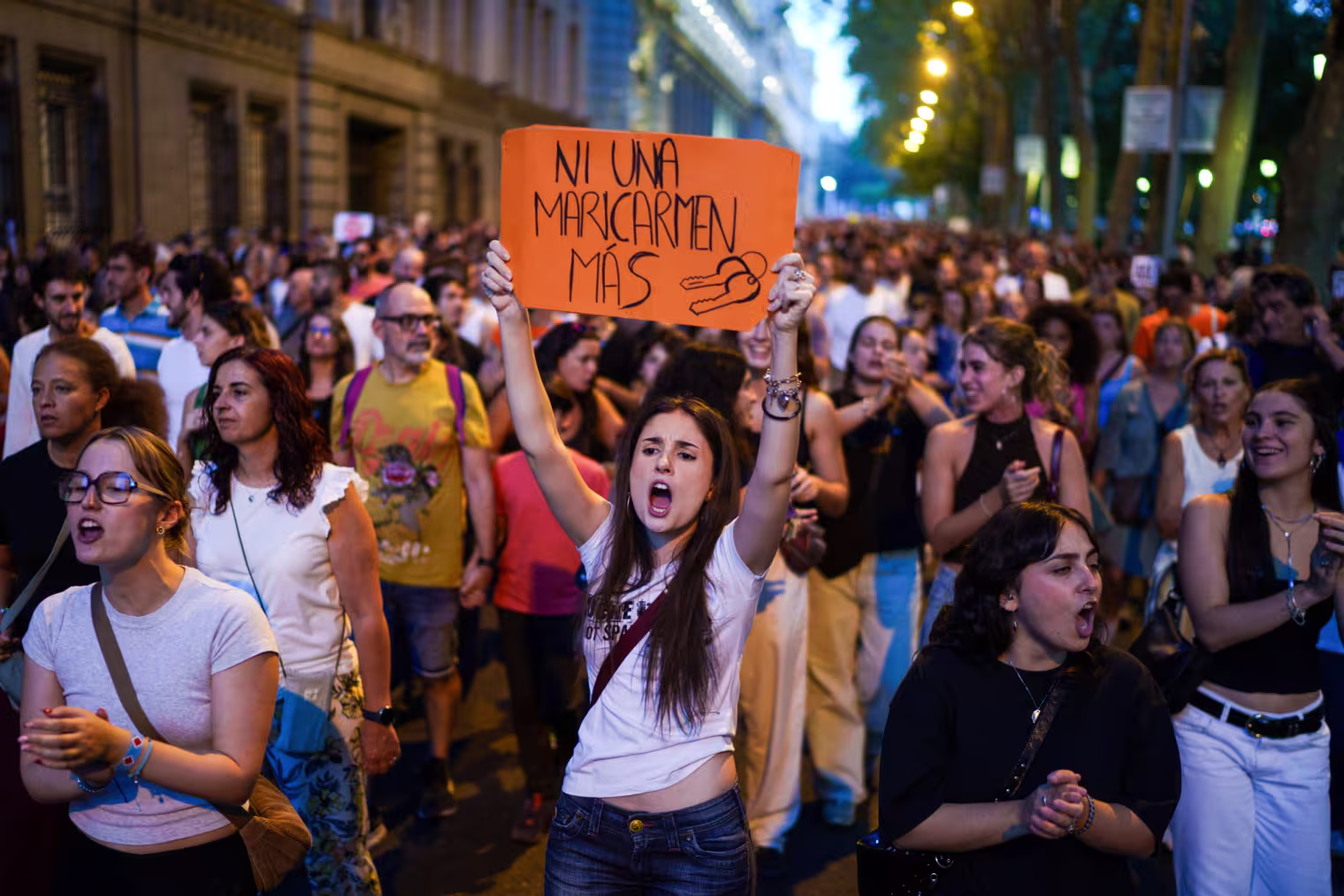 A woman holds an orange sign with black text in marker that reads "Ni Una Maricarmen Más" with drawn picture of keys