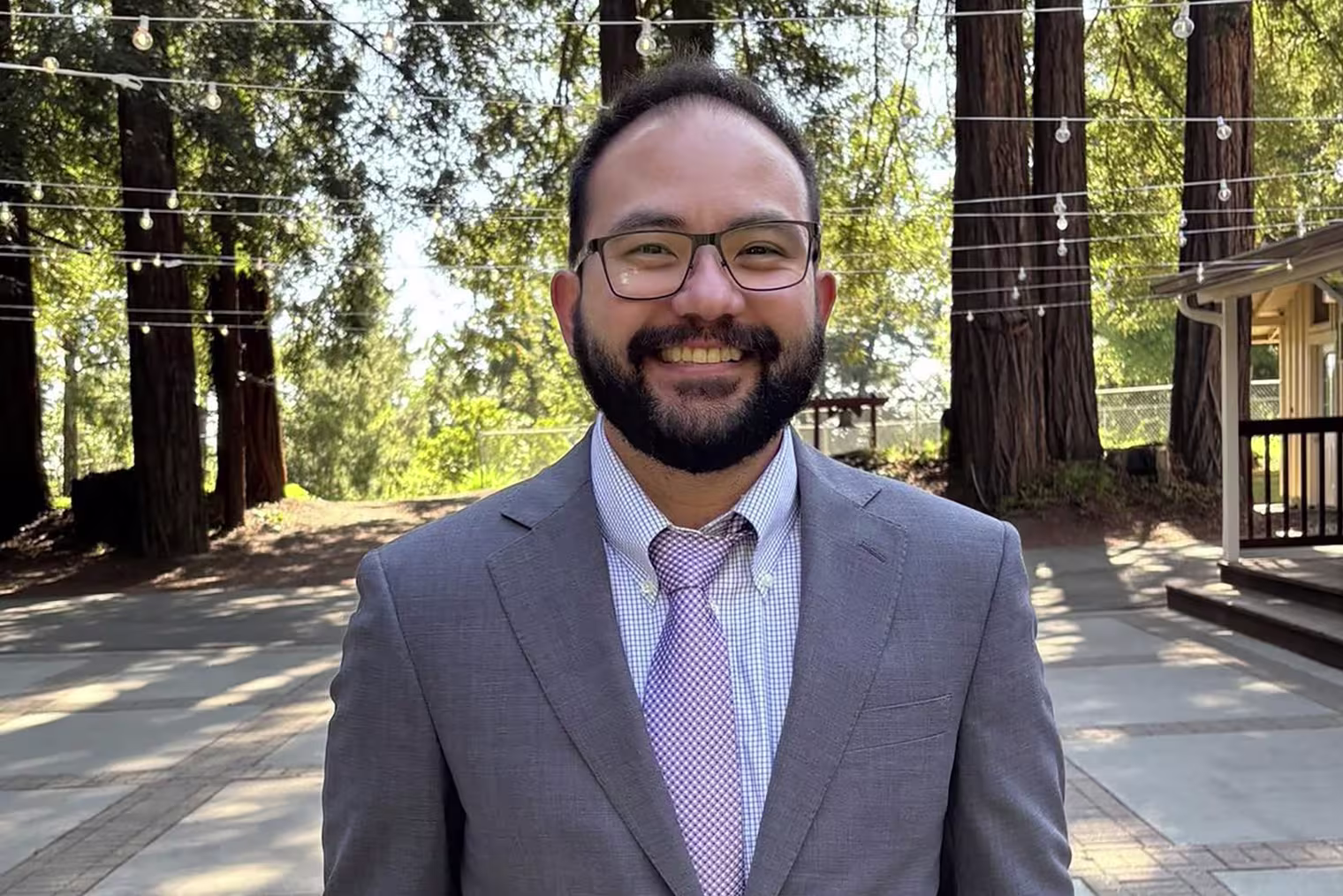 Image: Jonathan McKinsey wearing a suit, stood in front of trees draped with string lights.