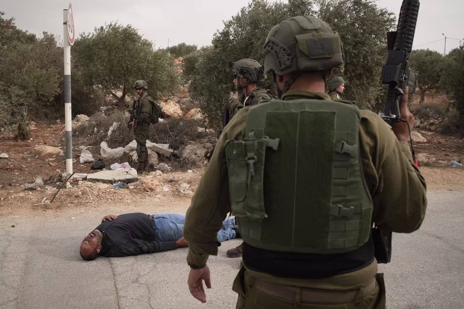Image: Israeli soldiers stand beside a wounded Palestinian man lying on a road, holding his chest with an expression of pain.
