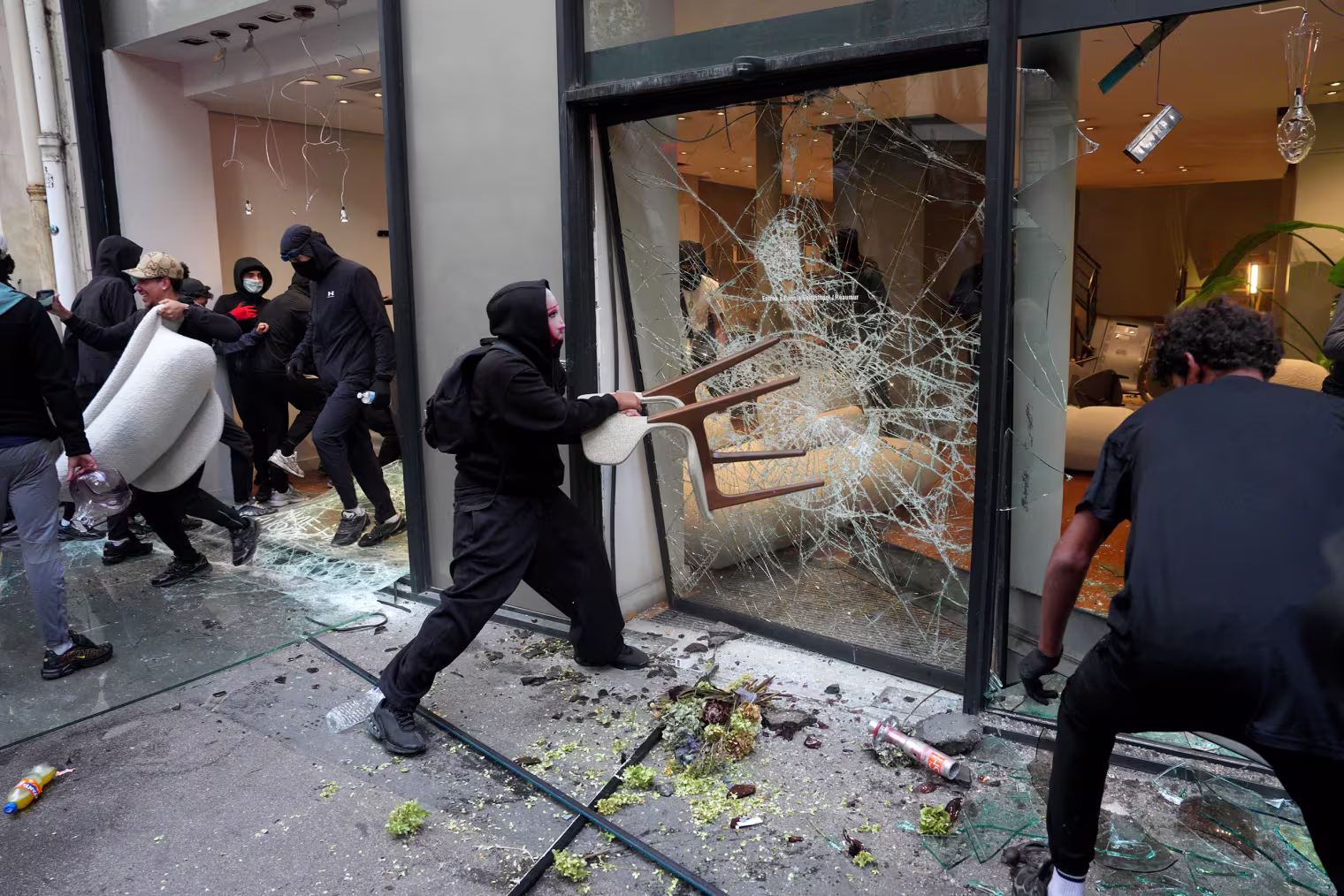 Image: A protesters smashes a retail store glass window with a chair.