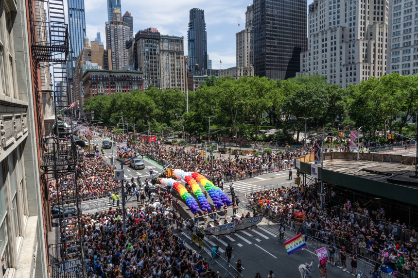 High-angle aerial view of a massive crowd at the NYC Pride 2025 parade on a city street lined with skyscrapers and trees. In the center of the parade route, participants march behind a large banner while holding giant, arched rainbow balloon installations above the street.