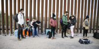 Asylum seekers from India, Cuba, and Colombia wait next to the U.S. border wall with Mexico, while being processed by U.S. border patrol in Yuma, Ariz., on Feb. 22, 2022.