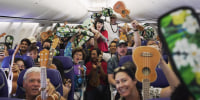 People on a plane hold their ukuleles up in the air, smiling for a camera.