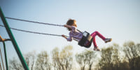 Little girl swings way up high into the sky above the trees on a swing set.