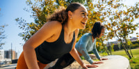 Cheerful women doing push-ups on retaining wall at park.