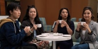 Four people holding white coffee cups look at something offscreen. They are sitting at a table on green couches.