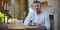 David Beckham in a white henley T-shirt with buttons and a wristwatch sits at a table and looks serious. 