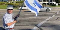 A man waves an Israeli flag in a park space along a city street