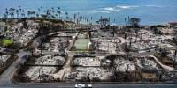 An aerial view of a recovery vehicle driving past burned structures and cars in Lahaina