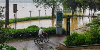 A cyclist peddles past flood waters near a river