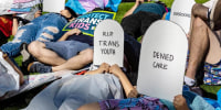 Protesters lie on the ground holding cardboard signs shaped like tombstones that read "Depression," "R.I.P. Trans Youth," "Denied Care" and "Overdosed"