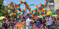 People walk through the streets during the Baltimore Pride Parade, balloons are shaped into the word "Love" in the background
