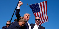 Donald Trump, with blood on his ear, raises up a fist while being escorted away by secret service outdoors, American flag flies in the background