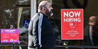 A pedestrian walks past a "Now Hiring" sign posted on a business storefront