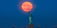 Full Harvest Moon Sets at Sunrise, the flame of the Statue of Liberty appears to touch the bottom of the moon in the sky