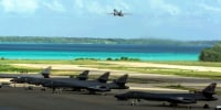 A plane takes off in the distance above a body of water, and five other planes are visible parked in the foreground.